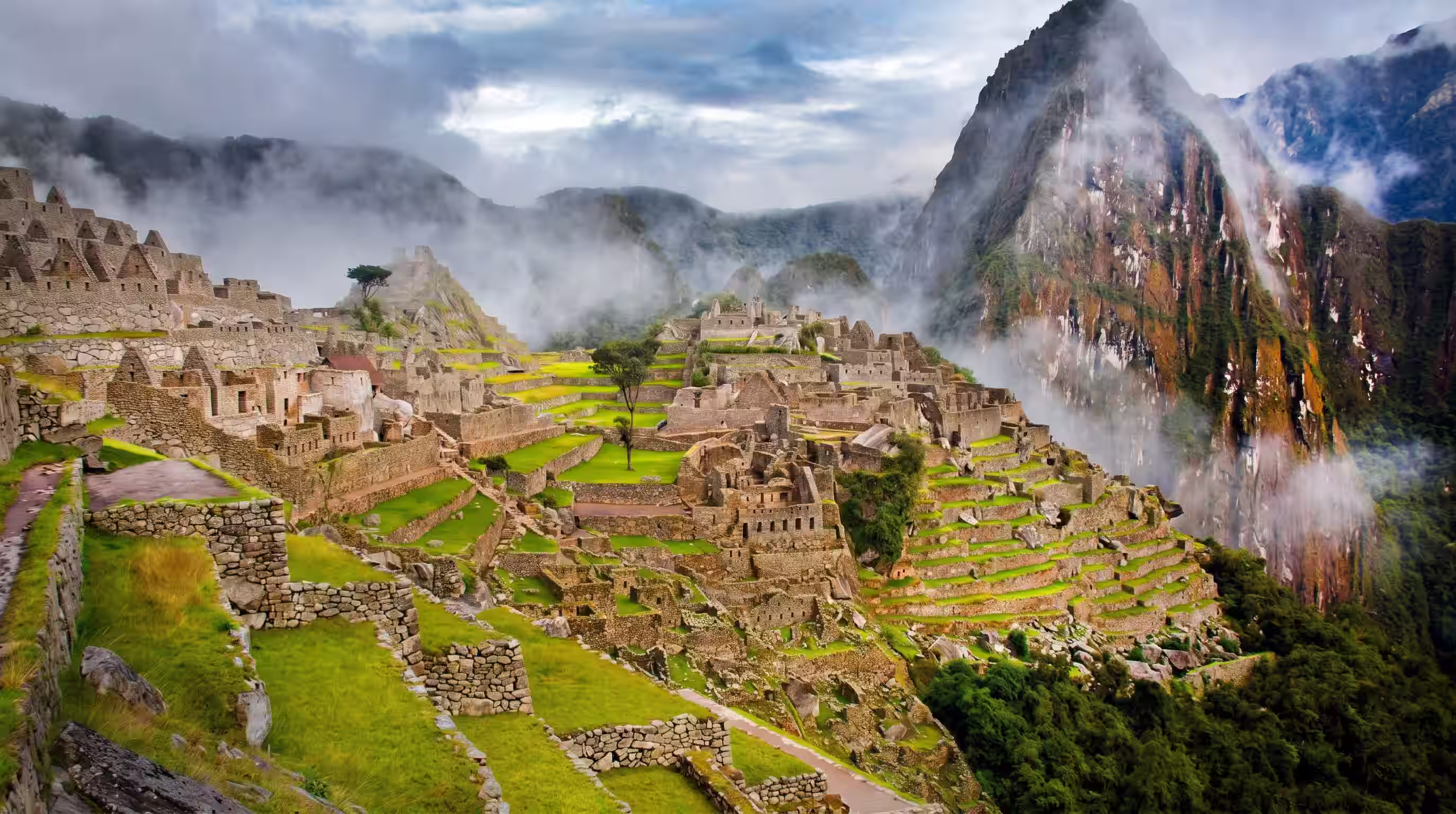 Terrazas y construcciones de Machu Picchu entre montañas, con nubes y neblina sobre las laderas.