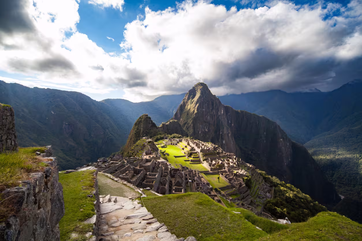 Vista de las terrazas y construcciones de Machu Picchu entre montañas, con un sendero de piedra en primer plano y nubes sobre el valle.